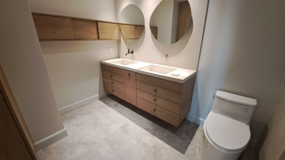 Salle de bain avec un meuble-lavabo en bois, deux miroirs ronds au-dessus, et des toilettes blanches à droite.