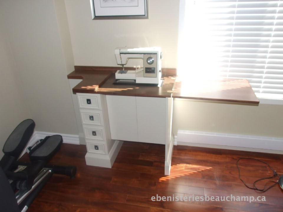 Une machine à coudre sur une table en bois avec tiroirs, près d'une fenêtre avec des stores, sur un sol en bois.