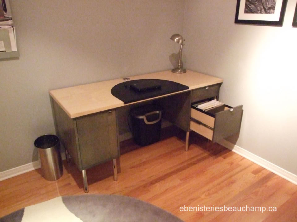 Bureau en bois clair avec tiroirs ouverts, lampe de bureau, poubelle en métal, et cadre photo sur le mur.