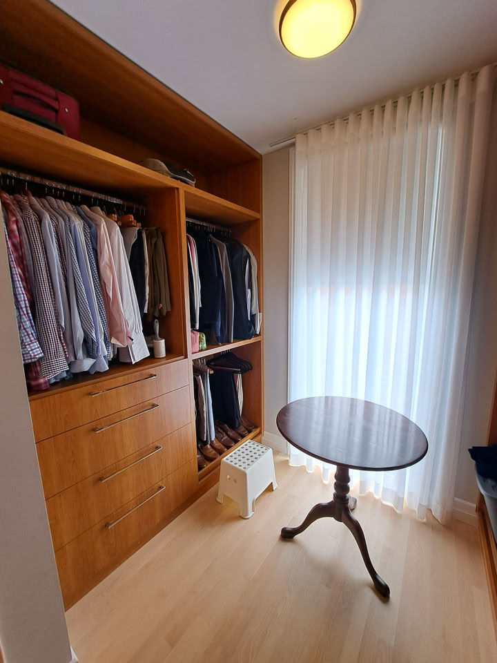 A walk-in closet with wooden shelves and drawers, clothes hanging on racks, a small round table, and a window with sheer curtains.