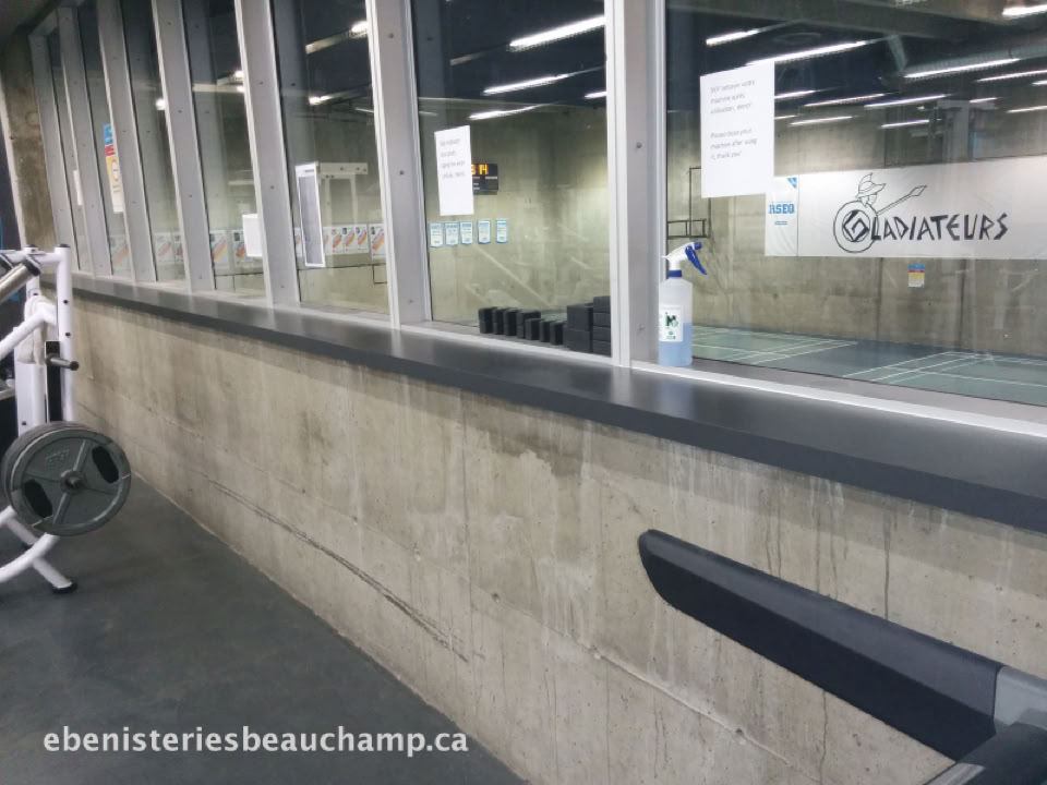 Salle de sport avec des équipements d'exercice, des fenêtres donnant sur un court de squash, et une bouteille d'eau posée sur le rebord.