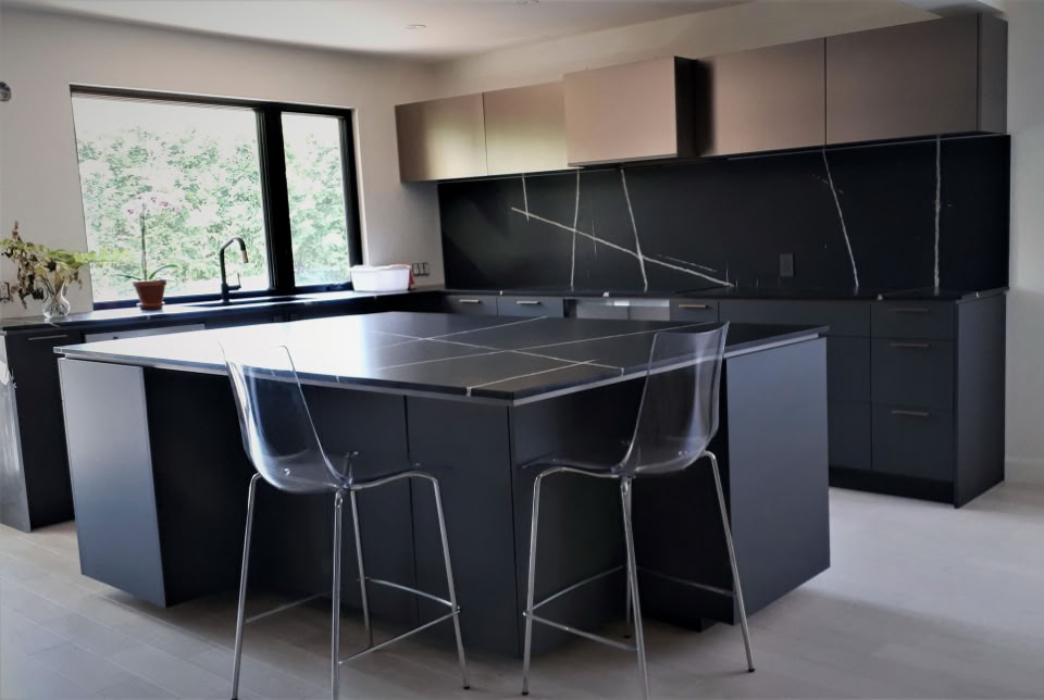 Modern kitchen with dark cabinetry, a large island with a dark countertop, two transparent bar stools, and a window with a view of greenery.