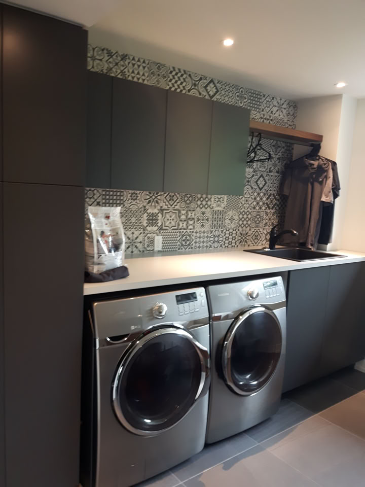 A laundry room with two front-loading washing machines, dark cabinets, a patterned tile backsplash, and a countertop with a small appliance.
