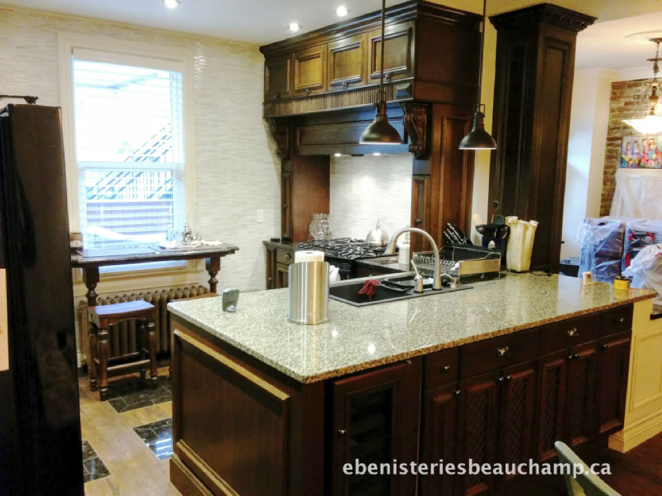 A kitchen with a large island featuring a granite countertop, a sink, and a paper towel holder. Dark wooden cabinets and a stove with an overhead range hood are in the background. A window is on the left, and a small table with chairs is near the window.