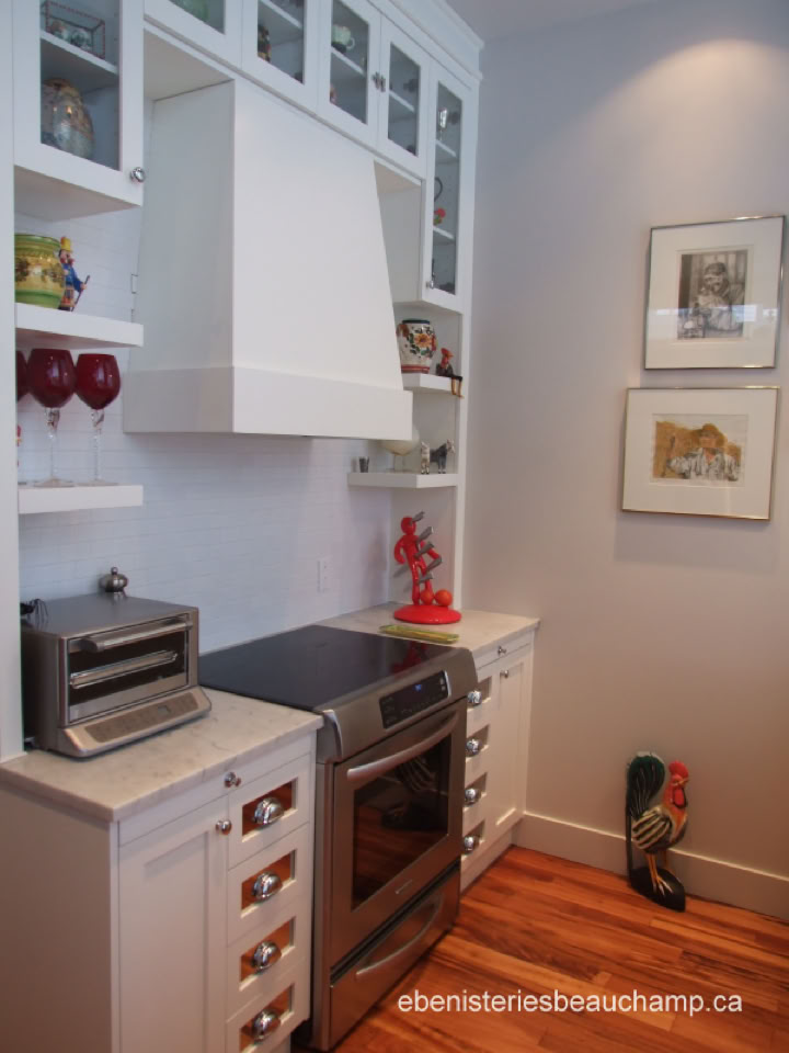 A kitchen corner with white cabinets, a stove, a range hood, a small countertop oven, and decorative items on shelves. Two framed pictures hang on the wall.