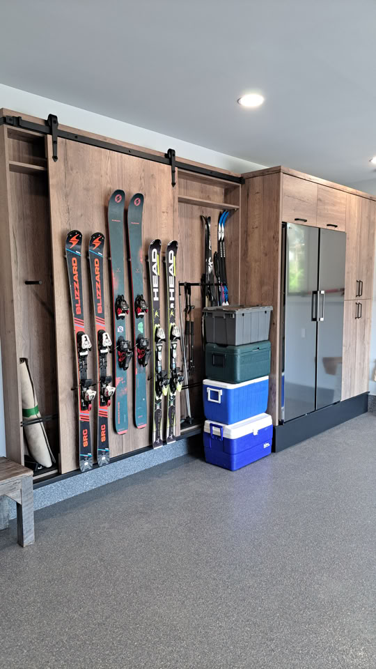 A garage with a wooden storage unit holding several pairs of skis and a baseball bat, alongside stacked blue and green storage bins.