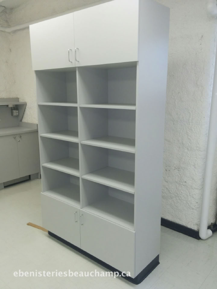 Grande étagère blanche avec plusieurs compartiments et deux portes en haut, située dans une pièce aux murs blancs.