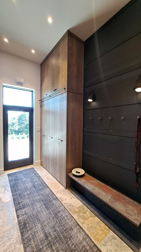A modern entryway with a tall wooden cabinet, a bench with a hat on it, dark wall panels with hooks, and a glass door leading outside.