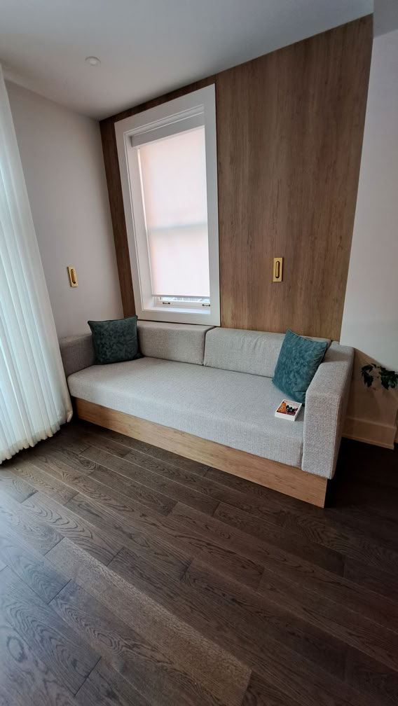 A modern room with a light gray sofa against a wooden accent wall, two teal cushions, a window with a sheer curtain, and dark wooden flooring.