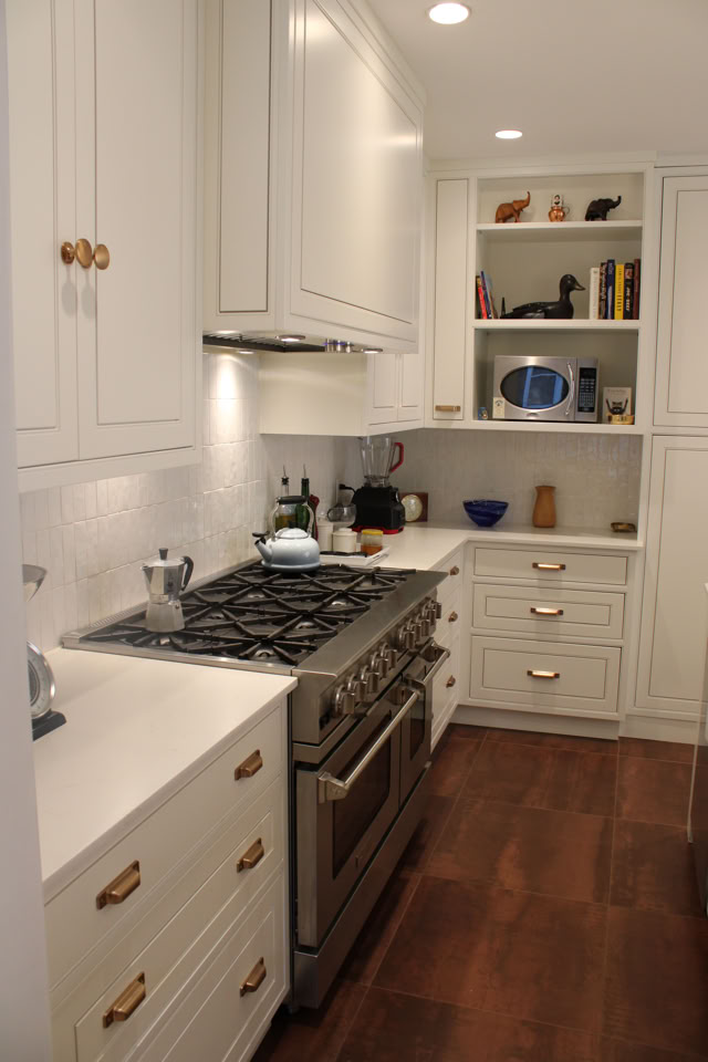 A narrow kitchen with white cabinets, a stainless steel stove, and wooden flooring. Shelves with books and decorative items are visible in the background.