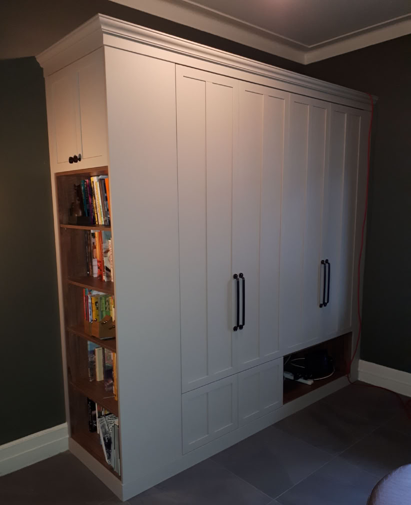 A white built-in wardrobe with two large handles, featuring a side bookshelf filled with books and a small open compartment at the bottom.