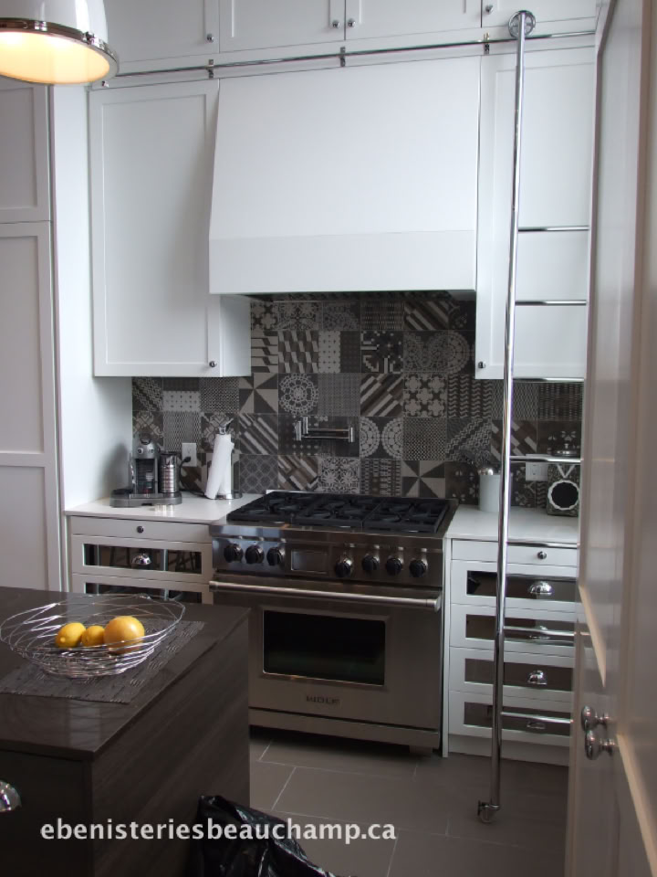 A modern kitchen with a stainless steel stove, patterned tile backsplash, white cabinets, and a hanging light fixture. A small island with a fruit bowl is in the foreground.