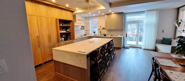Cuisine moderne avec un grand îlot central, des armoires en bois clair, un sol en bois foncé, et une table à manger avec des chaises. Fenêtres laissant entrer la lumière naturelle.