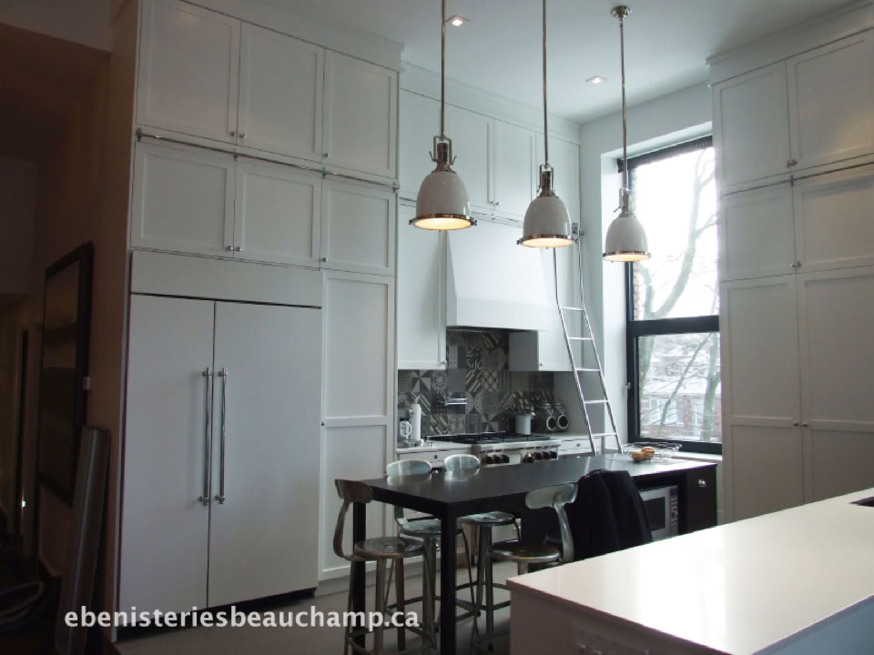 A modern kitchen with white cabinets, a large refrigerator, a central island with bar stools, pendant lights, and a window.