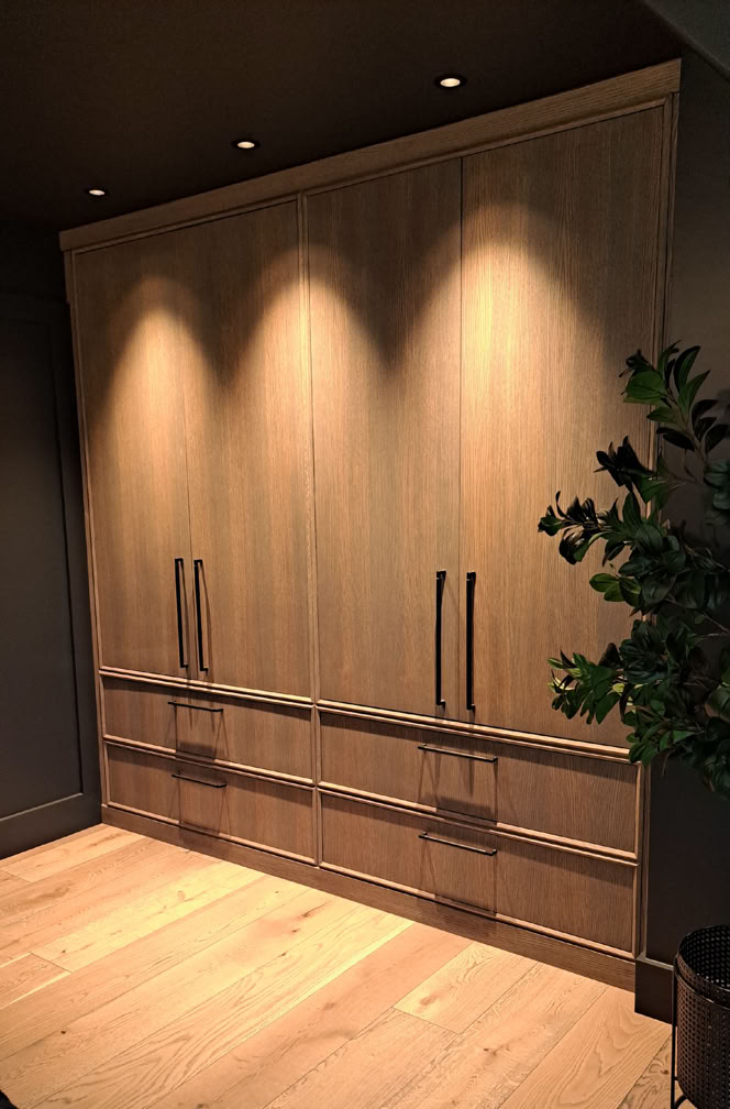 Wooden wardrobe with four vertical handles, illuminated by overhead lights, and a plant partially visible on the right.