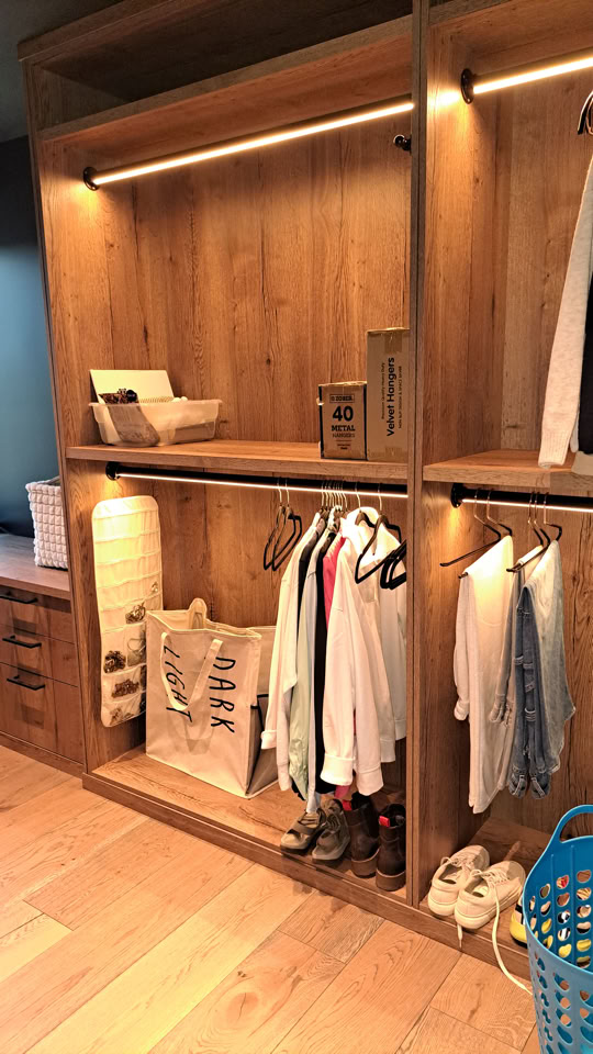 Wooden closet with shelves and hanging space, containing folded clothes, hanging garments, shoes, and decorative items.
