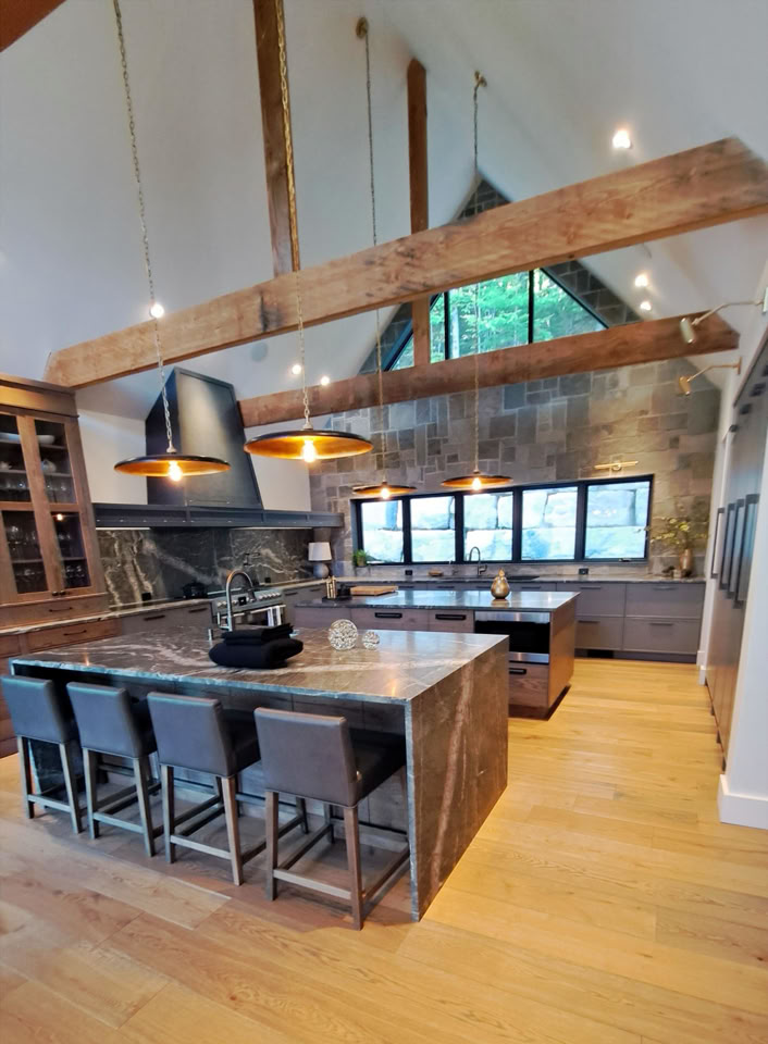 Spacious kitchen with wooden beams, large island with bar stools, pendant lights, and a stone wall with windows.