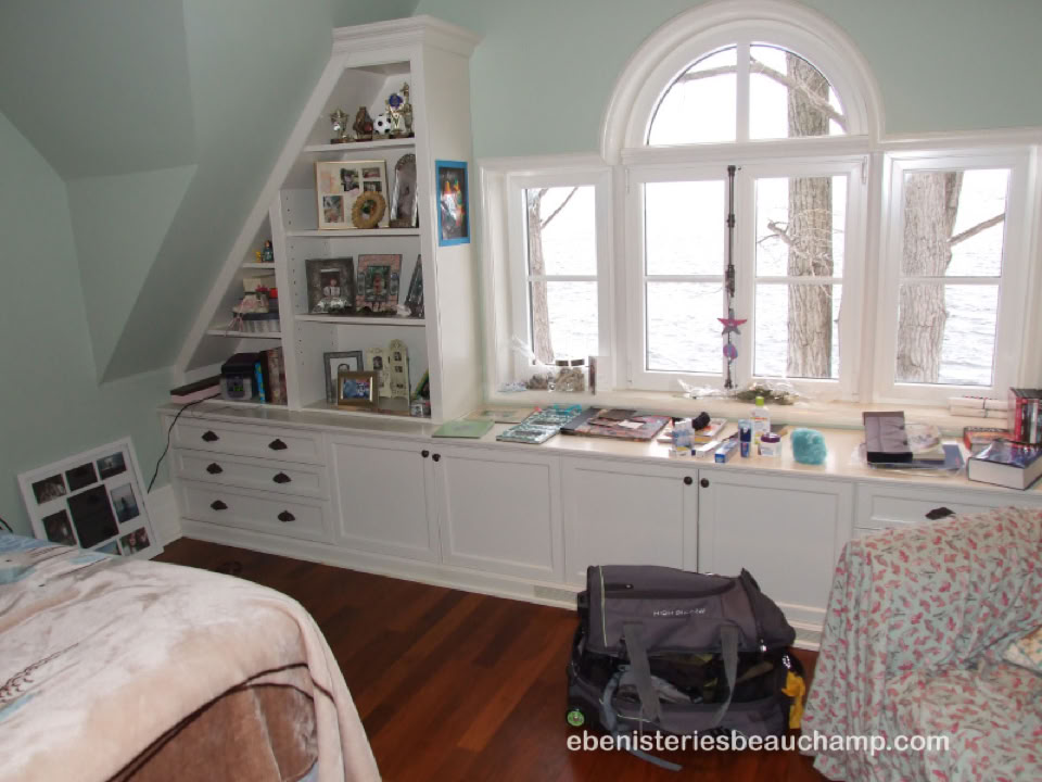 A cozy room with a bed, built-in shelves, and a window with a view of trees. The shelves contain books and decorative items. A bag is on the floor, and the walls are light-colored.