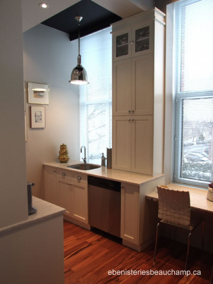A small kitchen with white cabinets, a stainless steel dishwasher, a sink under a window, a pendant light, and a wooden floor.