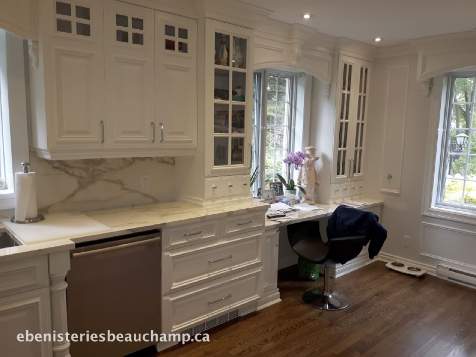 White kitchen with marble countertops, glass-front cabinets, a desk area with a chair, and a window with a view of greenery.