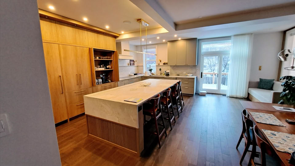 A modern kitchen with a large island, wooden cabinets, and a dining table. The room has recessed lighting, a window with sheer curtains, and a door leading outside.