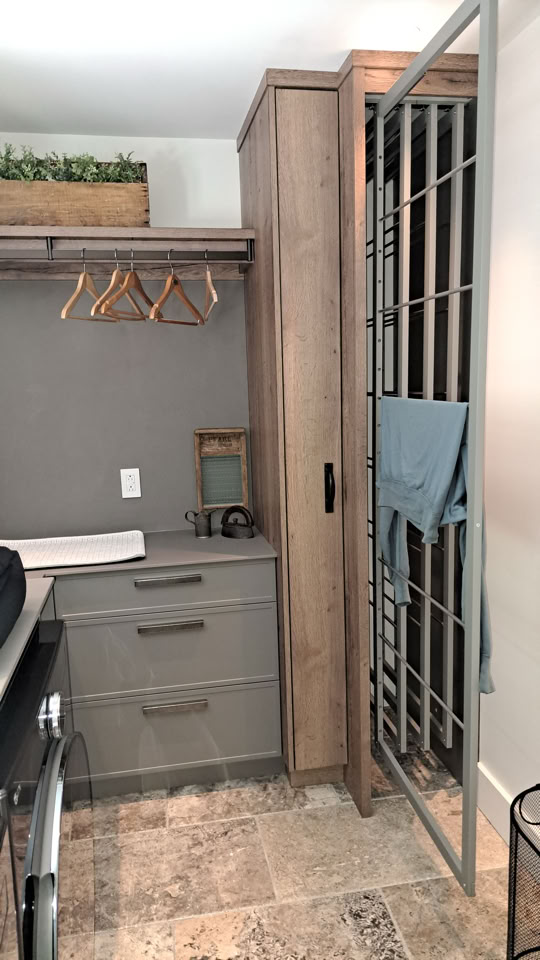 A laundry room with a washing machine, gray cabinets, a countertop, and a hanging rod with wooden hangers. A metal gate is open, and a towel is draped over it. A plant is on top of the cabinet.