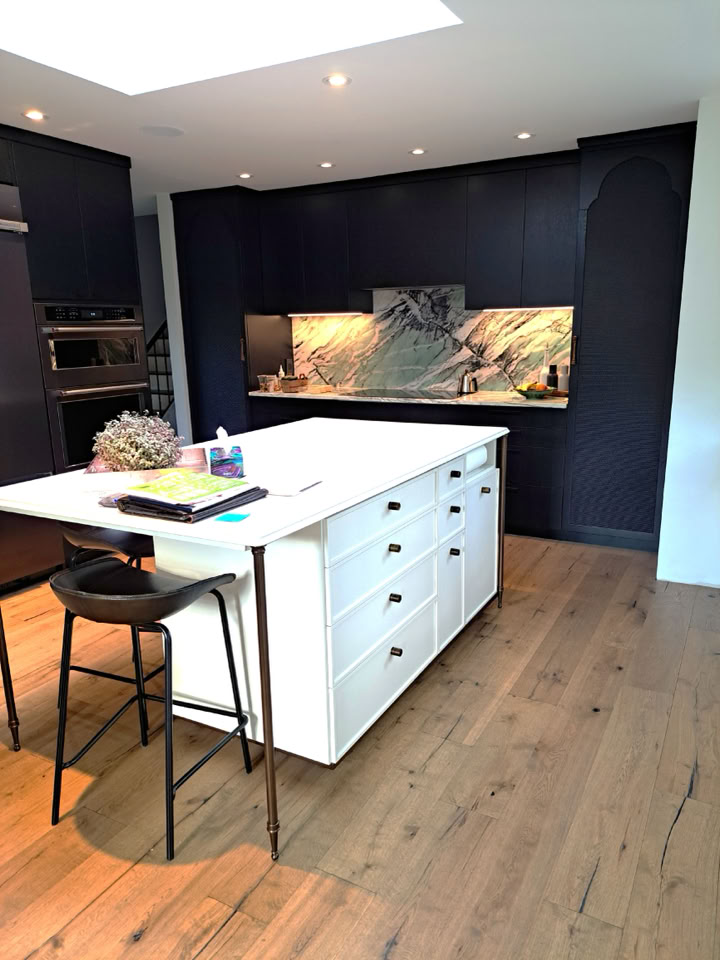 Modern kitchen with a white island, black bar stool, dark cabinets, marble backsplash, and wooden floor.