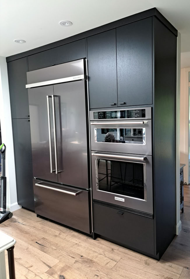 A modern kitchen with a large stainless steel refrigerator, built-in double ovens, and dark cabinetry.