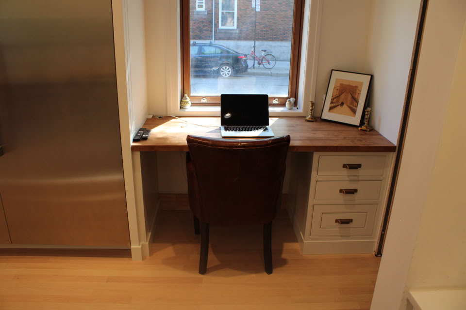 A small wooden desk with a laptop, a framed picture, and a chair, positioned in front of a window with a view of a street.