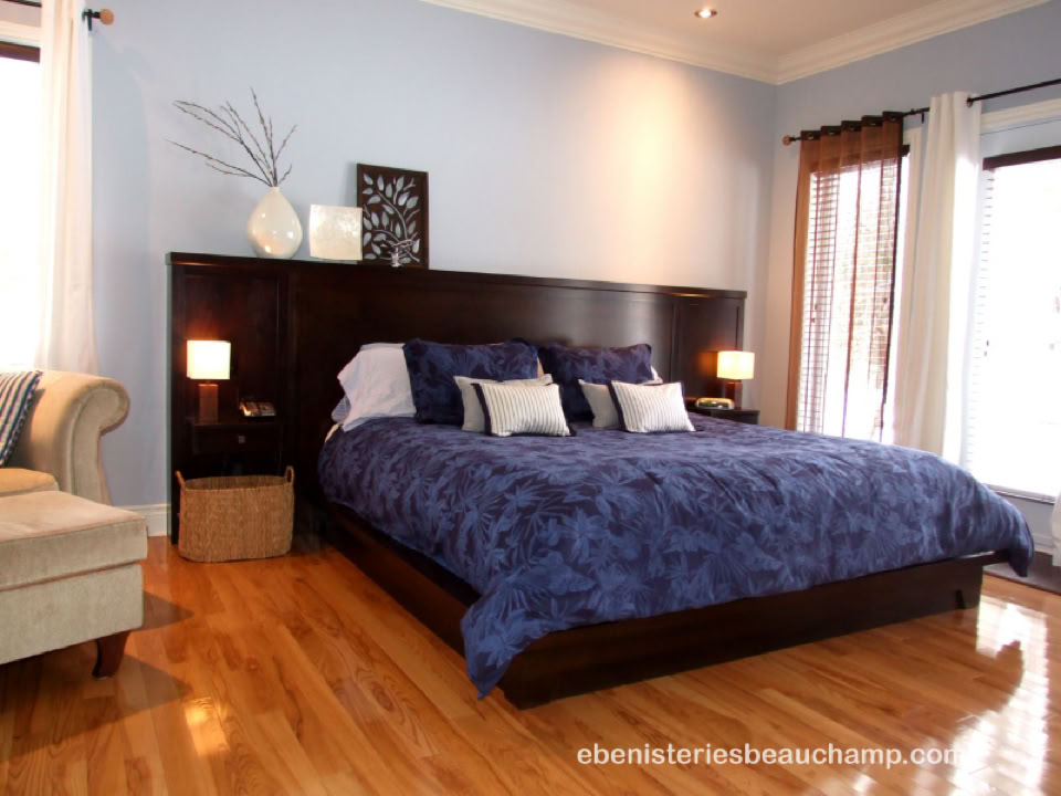 A bedroom with a large bed covered in a blue comforter, two bedside tables with lamps, a cushioned chair, and a window with curtains.