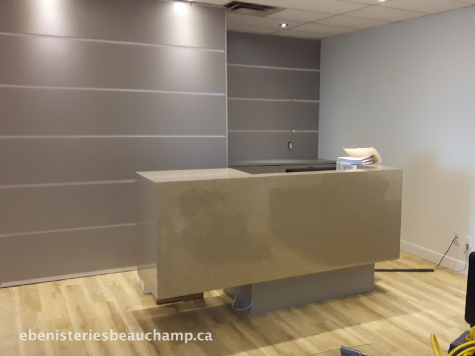 Bureau moderne avec un comptoir beige, sol en bois clair, murs gris et blancs, et plafond avec éclairage encastré.