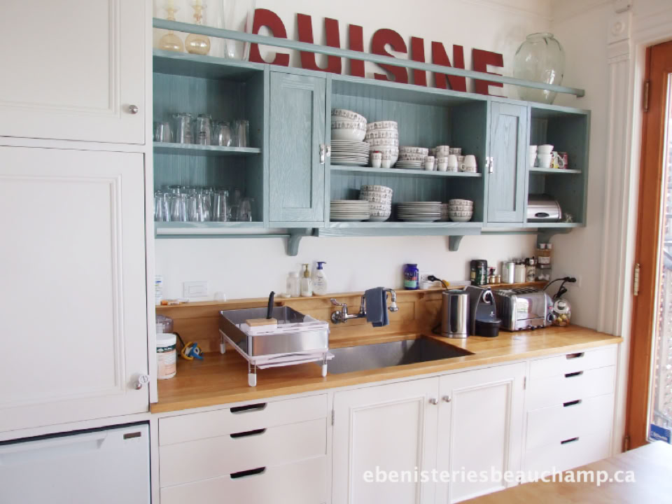 A kitchen with white cabinets, a wooden countertop, a sink, and various kitchen appliances. Open shelves display dishes and glasses. The word "CUISINE" is partially visible on the wall above the shelves.