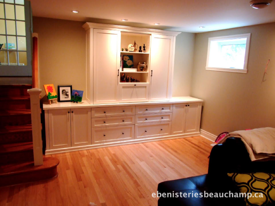 A room with wooden flooring, a white built-in cabinet with shelves and drawers, a small window, and a staircase on the left.